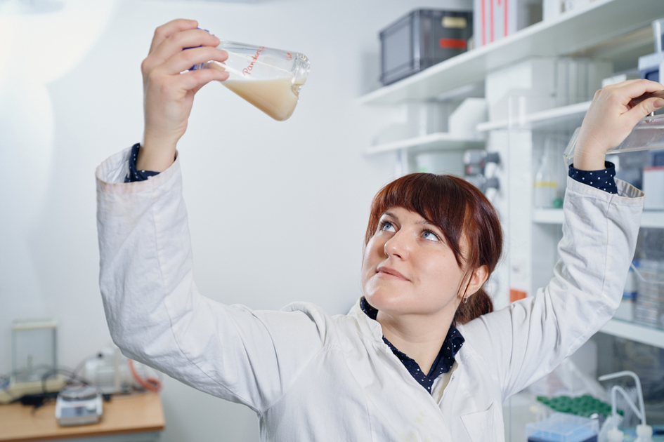 Linda Muskat im Porträt im Labor In der Hand hält sie einen Glaskolben mit einer milchig braunen Flüssigkeit