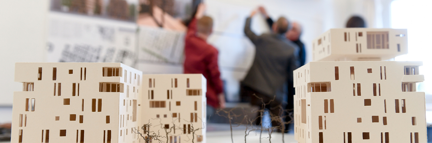 Architektur-Modelle sind im Vordergrund zu sehen, im Hintergrund zeigen Studierende auf Plänen, die an der Wand befestigt sind.