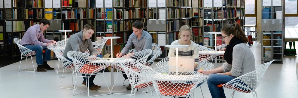 Studierende sitzen an Tischen und lernen. Im Hintergrund sind Bücherregale zu sehen.